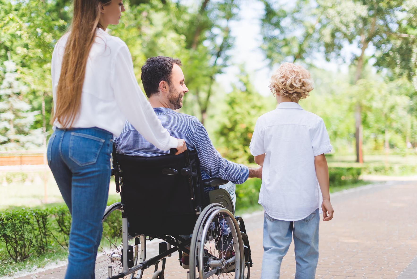 back view of mother rolling wheelchair with disabled father in park